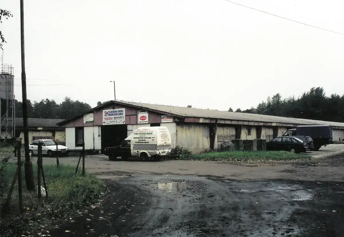 Stara hurtownia materiałów budowlanych z blaszanym dachem i samochodami dostawczymi przed wejściem.