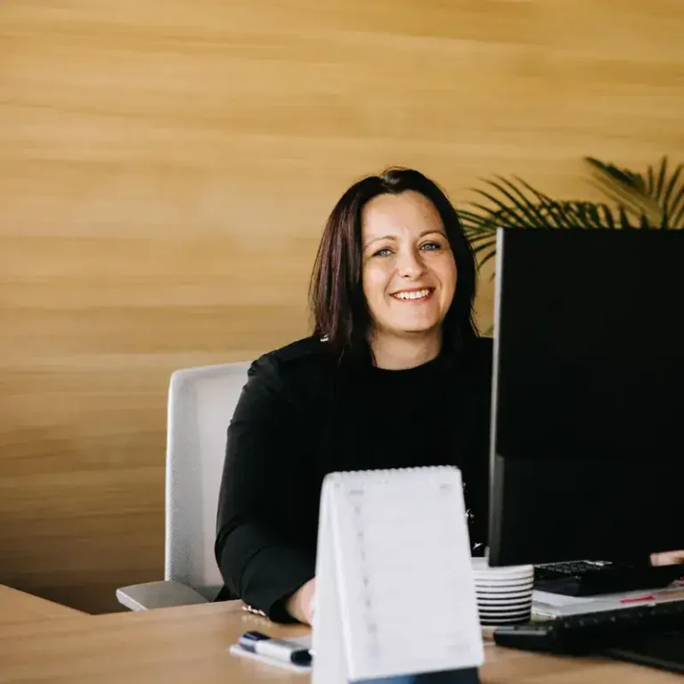 Pracowniczka EKOBau siedząca przy biurku, uśmiechnięta, obsługująca klientów i udzielająca doradztwa budowlanego w nowoczesnym biurze.