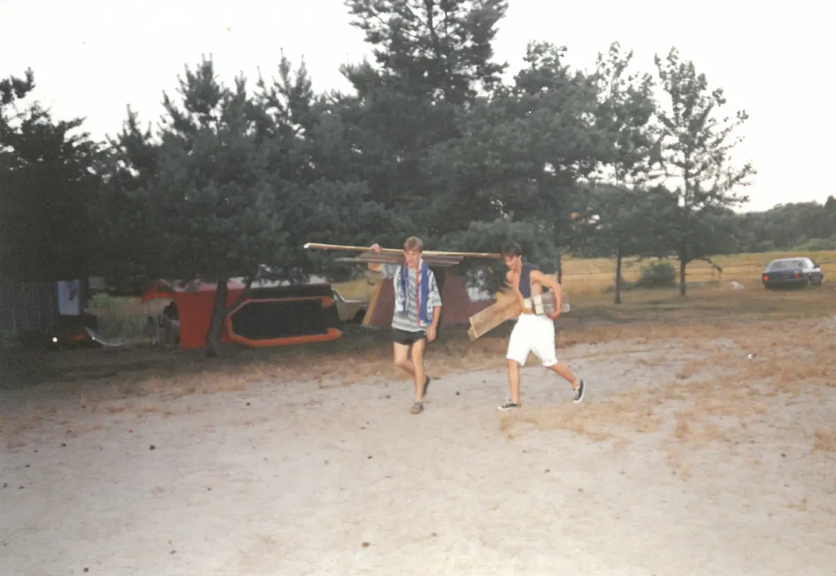 Dwóch młodych mężczyzn niosących deski i rury na tle drzew i namiotów w letnim krajobrazie.