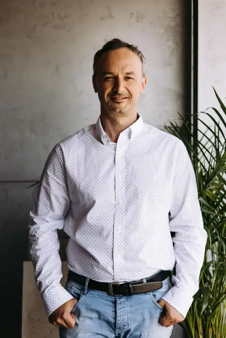 Mężczyzna w białej koszuli stojący w biurze EKOBau, uśmiechnięty, symbolizujący nowoczesne podejście firmy do branży budowlanej.