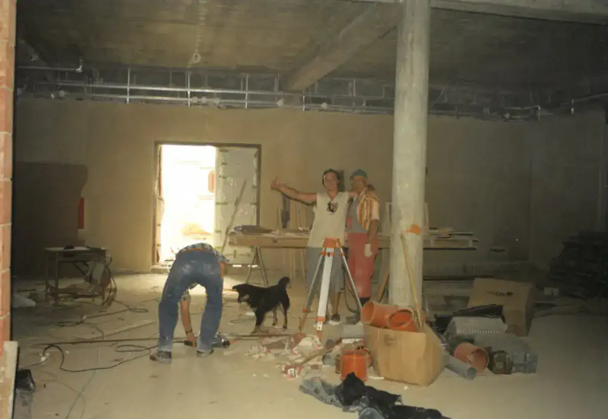 Grupa pracowników w trakcie remontu – wnętrze w stanie surowym, sprzęt budowlany i pies w tle.